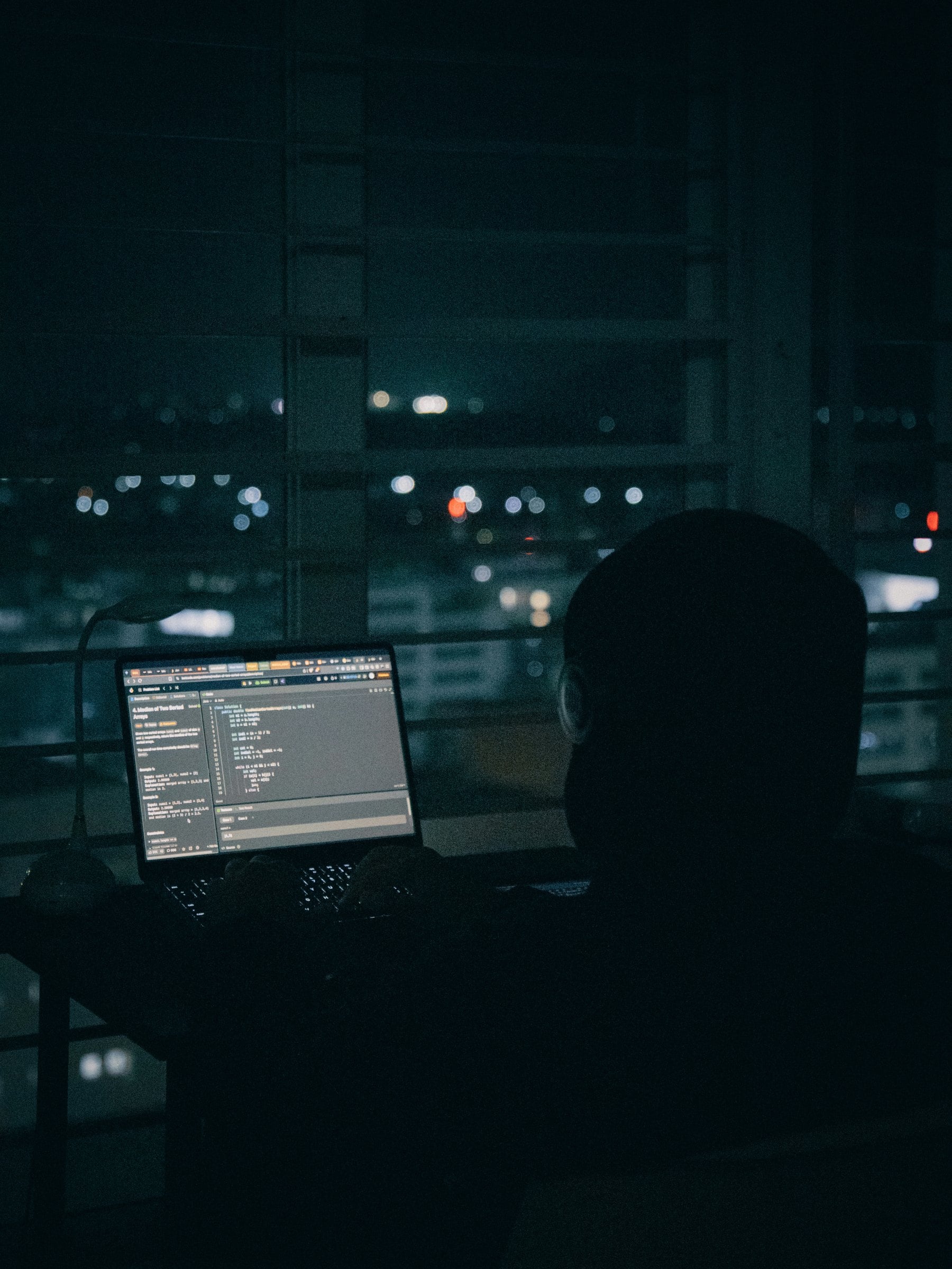 夜景の見える暗い部屋で、ノートPCのコードに向かう人の後ろ姿